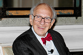Eric R Kandel, sitting looking to the side, smiling with a red bow tie, white shirt and black coat. 