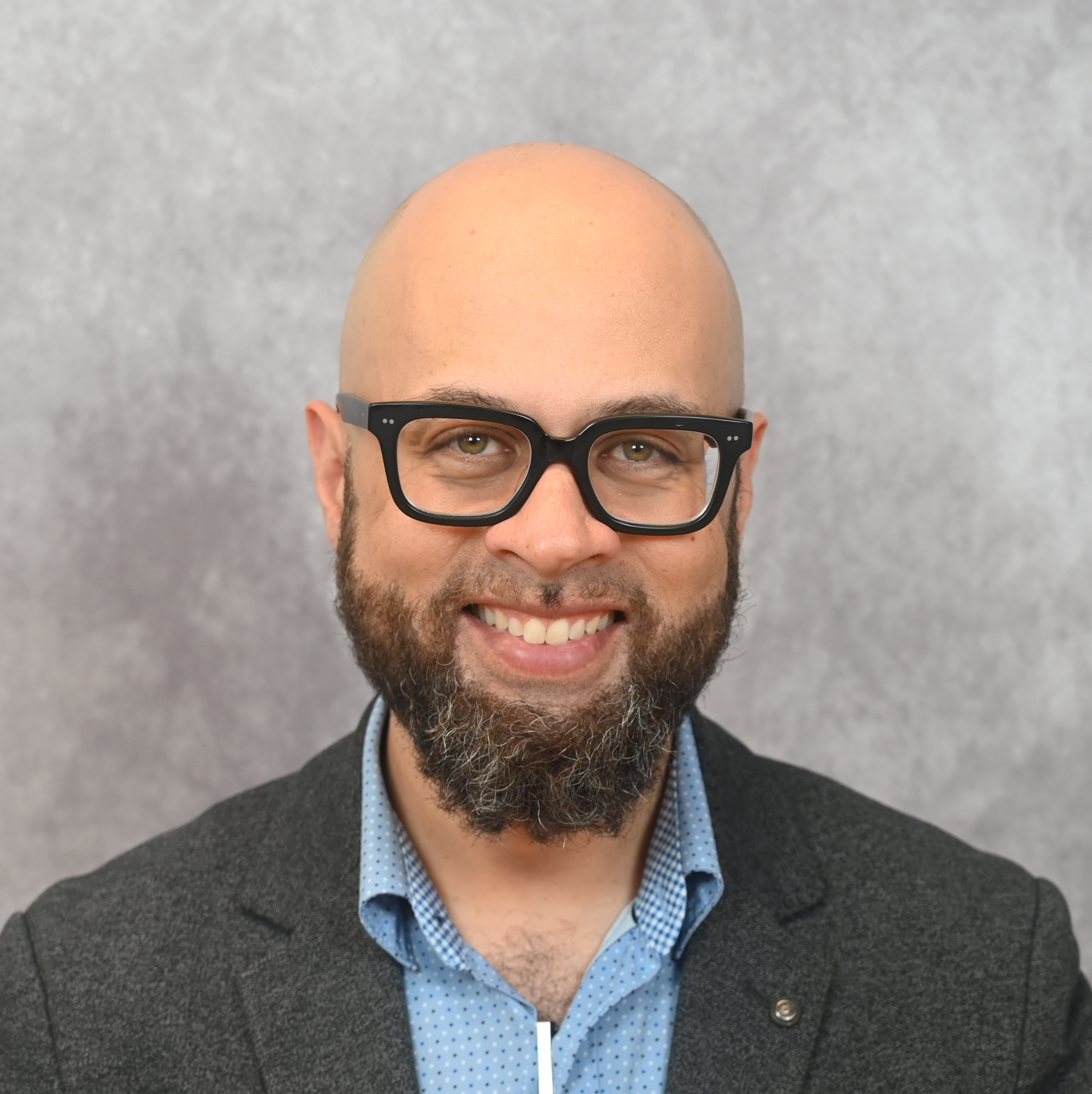 Headshot of a smiling bald man with a full beard and mustache, wearing black rectangular glasses, a light blue patterned button-up shirt, and a dark blazer, posed in front of a light gray mottled background.