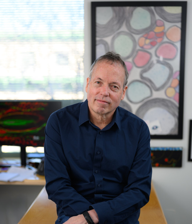A middle‑aged man with short gray hair and a navy button‑down shirt sits on the edge of a wooden desk in an office, looking at the camera with a slight smile. Behind him are large abstract artworks in black frames and a computer monitor displaying colorful graphics. Daylight filters in through a window with blinds, softly lighting the room.