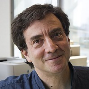 Middle‑aged person with short, dark, wavy hair smiles softly at the camera. They are wearing a dark blue shirt, and the background shows a bright room with large windows and blurred office equipment.”