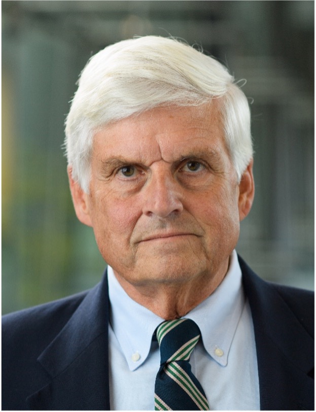 Older man with short white hair and light skin wearing a dark suit jacket, light blue dress shirt, and striped green and navy tie, looking directly at the camera with a neutral expression; soft, blurred indoor background.