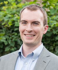 Portrait of a smiling man with short light brown hair, wearing a gray blazer and light blue shirt in front of leafy greenery.