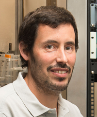 Portrait of a smiling man with short dark hair and a trimmed beard, wearing a light-colored collared shirt in a laboratory setting.