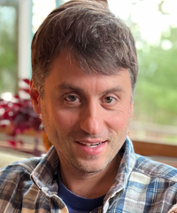 Portrait of a smiling man with short gray-brown hair, wearing a plaid shirt near a window.