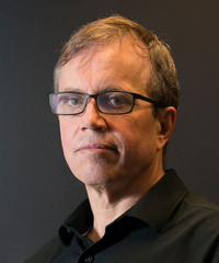 Portrait of a man with short light brown hair and rectangular glasses, wearing a black collared shirt against a dark background.