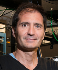 Portrait of a man with short dark hair, wearing a black shirt over a blue undershirt in a technical workspace.