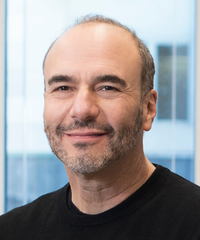 Portrait of a smiling man with short hair and a trimmed beard, wearing a black shirt against a softly blurred windowed background.