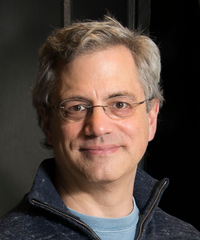 Portrait of a man with gray-brown hair and glasses, wearing a dark zip-up sweater over a blue shirt against a dark background.