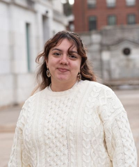 Portrait of a smiling woman with brown hair, wearing a white cable-knit sweater outdoors in front of historic buildings.