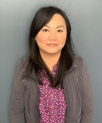 A woman with long dark hair wearing a gray cardigan over a patterned burgundy blouse, standing against a gray background.