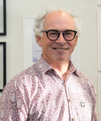 Portrait of a smiling man with white hair and round glasses, wearing a pink patterned shirt against a light-colored wall.