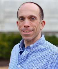 A man with a shaved head wearing a light blue checkered shirt, standing outdoors with blurred greenery and a building in the background.
