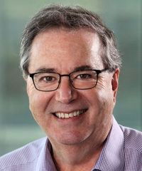 Portrait of a smiling middle-aged man with short gray hair and rectangular glasses, wearing a purple checkered shirt against a softly blurred background.