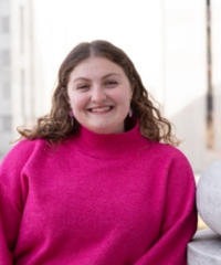 Portrait of a smiling woman with shoulder-length curly brown hair, wearing a bright pink turtleneck sweater.