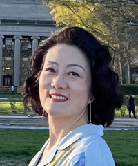 A smiling woman with short dark hair wearing a light-colored blouse, photographed outdoors with a university building, lawn, and trees in the background.