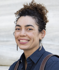 Portrait of a smiling woman with curly dark hair in a bun, wearing a navy patterned shirt, earrings, and a shoulder bag.
