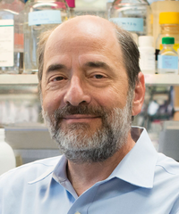 Portrait of a smiling man with short brown-and-gray hair and a salt-and-pepper beard, wearing a light blue collared shirt in a laboratory or pharmacy setting.