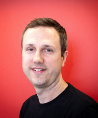 Portrait of a man with short brown hair, wearing a black shirt against a bright red background.