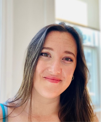 Portrait of a smiling woman with long brown hair, wearing a blue top, against a softly lit indoor background.