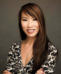 Portrait of a smiling woman with long dark hair, wearing a patterned blouse and necklace against a dark background.
