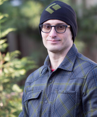A person wearing glasses, a dark beanie, and a blue-gray plaid shirt, standing outdoors in front of blurred greenery.