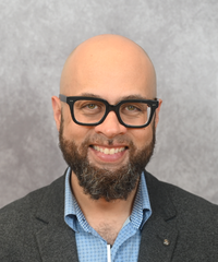 Smiling bald man with a full dark beard and black-rimmed glasses, wearing a gray blazer over a light blue collared shirt, posed in front of a mottled gray backdrop.
