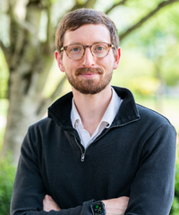 A man with short brown hair, a beard, and round glasses stands outdoors with his arms crossed, wearing a dark quarter‑zip sweater, a white collared shirt, and a smartwatch, with soft green trees and foliage blurred in the background.