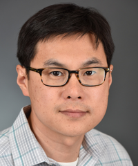 Head-and-shoulders portrait of a man wearing black rectangular glasses and a light plaid collared shirt against a gray background.