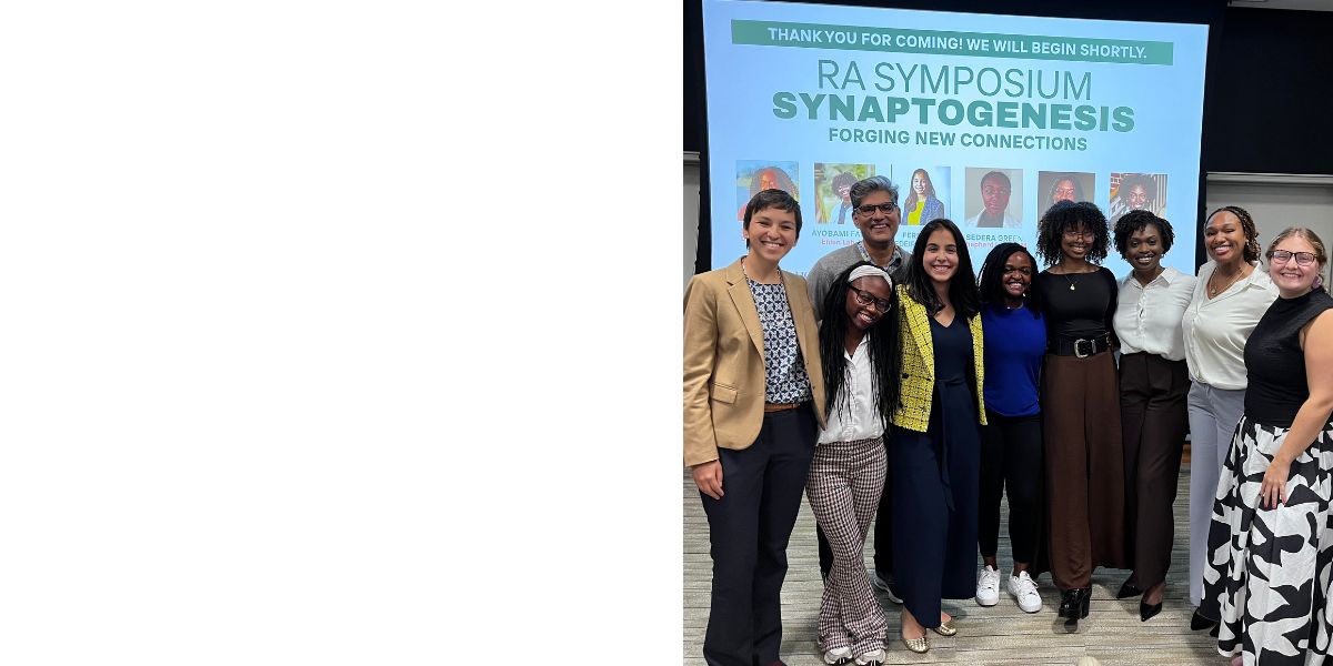 A group photo of nine people posing together at the RA Symposium "Synaptogenesis: Forging New Connections." They are standing in front of a large presentation screen with event details and speaker photos. The room features conference-style seating and a wooden podium in the background.