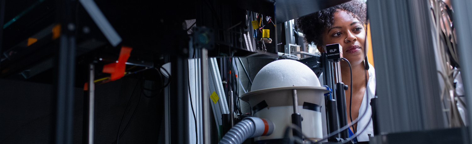 Scientist examining complex laboratory equipment with cables, sensors, and a white spherical component. The researcher, wearing a lab coat, looks focused as she observes the setup in a dimly lit lab environment.