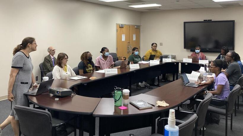 A group of people sits around a large, U-shaped table in a classroom setting, engaged in a discussion. Some participants have laptops, notebooks, or drinks in front of them, and a few are wearing masks. At the front of the room, Dr. Roz Segal (HMS) stands near a laptop, appearing to lead or present to the group. The walls are plain, and a large monitor is mounted on the wall behind the participants.