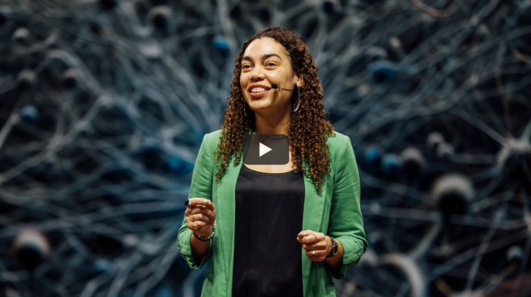 A woman with curly hair, wearing a green blazer and a black top, stands on stage smiling and speaking with a headset microphone. The background features an abstract design resembling interconnected neural or network pathways. There is a play button overlay in the center, indicating this is a video thumbnail.