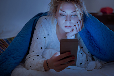 a girl laying on her stomach in bed, under the a blue blanket looking at her phone in a dimmed setting the light is illuminating from the phone. 